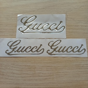 Może przedstawiać: Trzy srebrne metaliczne naklejki z napisem "Gucci" napisanym kursywą.