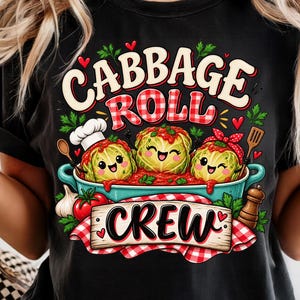 Op de afbeelding: Een zwart t-shirt met een grafische print van drie getekende koolrolletjes in een blauwe pan. De tekst luidt "CABBAGE ROLL CREW" in gestileerde letters. Het ontwerp bevat kleine rode hartjes, een koksmuts en een rood geruite doek.