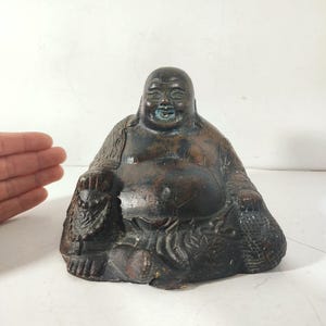 May include: A dark brown, seated statue of a smiling, chubby Buddha. The statue is holding a bag in one hand and a small figure in the other.