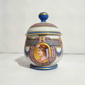 Peut inclure: Un pot en porcelaine blanche avec un couvercle bleu et un motif or et rose. Le pot présente un portrait d'une femme avec une couronne florale et un voile.