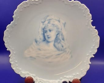 May include: A white porcelain plate with a scalloped edge features a blue and white image of a woman's portrait. The plate is on a blue background.