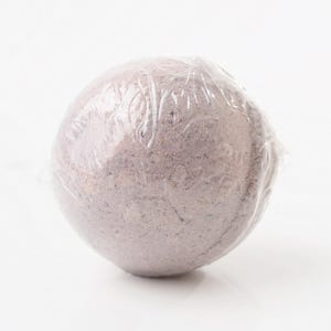 May include: A round, lavender-colored bath bomb wrapped in clear plastic. The bath bomb has a textured surface and appears to be made of a granular material. The packaging is transparent, allowing the product to be visible.