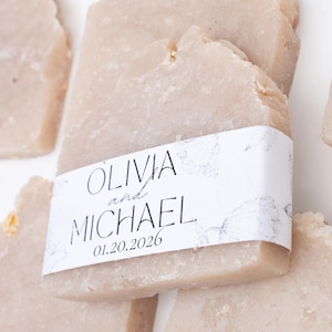 May include: Several light beige soap bars with rough edges are arranged on a white surface. One bar has a white label with the names "OLIVIA and MICHAEL" and the date "01.20.2026" printed on it. The soap bars appear to be handmade.
