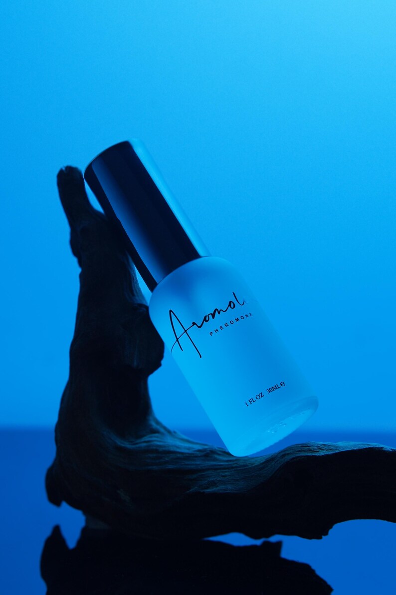 Puede incluir: Una botella de vidrio esmerilado azul claro con tapa negra, etiquetada "Aromol Pheromone" en escritura cursiva. La botella es de 30 ml y est&aacute; colocada sobre un fondo azul y un elemento de madera oscura texturizada.