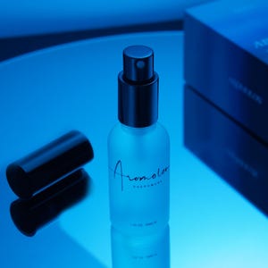 Puede incluir: Un frasco de perfume de vidrio esmerilado con tapa negra y la palabra "Aromolor" en cursiva. El frasco contiene 30 ml y est&aacute; sobre una superficie reflectante, con una caja oscura al fondo. La imagen est&aacute; ba&ntilde;ada en luz azul.