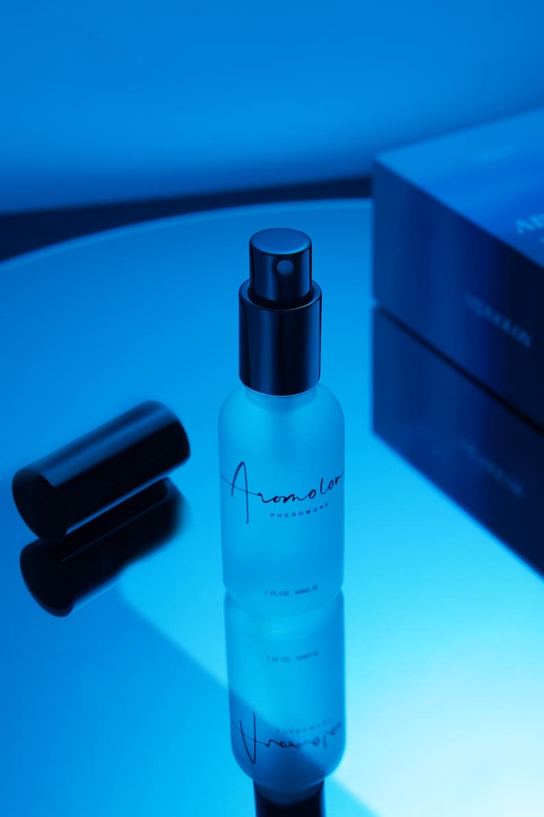 Puede incluir: Un frasco de perfume de vidrio esmerilado con tapa negra y la palabra "Aromolor" en cursiva. El frasco contiene 30 ml y est&aacute; sobre una superficie reflectante, con una caja oscura al fondo. La imagen est&aacute; ba&ntilde;ada en luz azul.