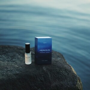 Puede incluir: Un frasco de colonia de vidrio transparente con tapa negra y una caja a juego. La caja es azul oscuro con la palabra "MEN" en la parte superior y "AROMOLON PHEROMONE COLOGNE" impresa en la parte delantera. El conjunto est&aacute; sobre una roca gris oscura.