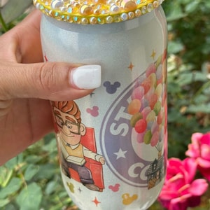 May include: White travel mug with a sparkly finish and a colorful Up movie design featuring Carl Fredricksen and balloons. The lid is decorated with a yellow circle of iridescent rhinestones.