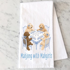 May include: A white kitchen towel featuring a printed illustration of four golden and brown doodle dogs sitting on light blue chairs around a table playing mahjong. The text "Mahjong with Mahgirls" is printed in light blue below the table.