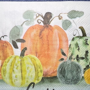 May include: A white paper napkin featuring a watercolor illustration of pumpkins in shades of orange, green, and yellow. The word "Gather" is written in black script across the bottom. The napkin has a textured surface.