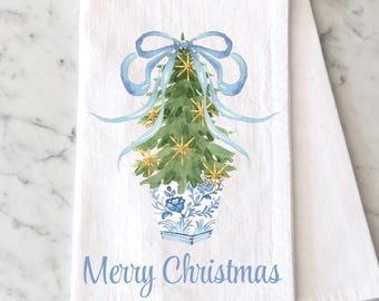 May include: A white kitchen towel featuring a watercolor illustration of a green Christmas tree in a blue and white floral pot. The tree is topped with a light blue bow and gold star ornaments. The text Merry Christmas is printed in blue below.