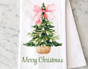May include: A white kitchen towel featuring a watercolor illustration of a green Christmas tree in a tan pot, topped with a large pink bow and decorated with small white ornaments. The text Merry Christmas is printed in green below the tree.