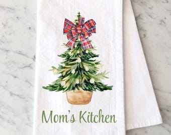 May include: A white kitchen towel featuring a watercolor illustration of a green Christmas tree in a tan pot, topped with a red and blue plaid bow. The tree is decorated with small white ornaments. The text Mom's Kitchen is printed in green.
