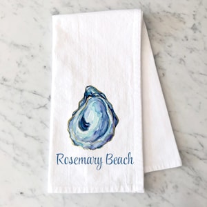 May include: A white kitchen towel featuring a watercolor illustration of a blue and purple oyster shell with gold edges. Below the shell, the text Rosemary Beach is printed in a blue script font. The towel is folded vertically on a white surface.