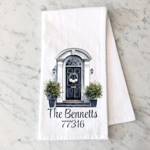 May include: White kitchen towel featuring a watercolor illustration of a black front door with a white wreath, flanked by potted plants. The text "The Bennetts" and the number "77316" are printed below the door.