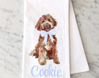 May include: A white kitchen towel featuring a watercolor illustration of a brown curly-haired dog wearing a light blue and white gingham bow. The name Cookie is printed in light blue script lettering below the dog illustration on the fabric.