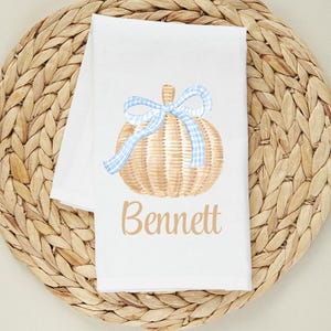 May include: White kitchen towel featuring a watercolor pumpkin design with a blue gingham bow. The pumpkin is tan with vertical lines. The name "Bennett" is written in tan script below the pumpkin.