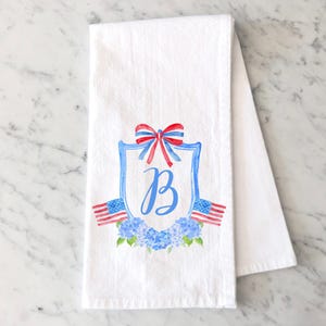 May include: White kitchen towel with a red, white, and blue bow, a blue shield with the letter "B" in white, and a wreath of blue hydrangeas. The shield is flanked by two American flags.