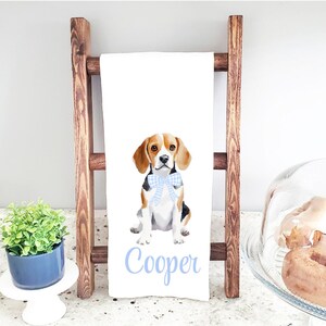 May include: White kitchen towel featuring a watercolor illustration of a beagle wearing a blue gingham bow tie. The dog's name, "Cooper," is written in light blue script below the image. The towel is displayed on a wooden ladder.