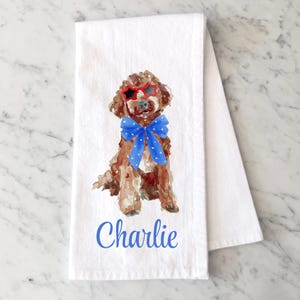 May include: A white kitchen towel featuring a watercolor illustration of a brown dog wearing red star-shaped sunglasses and a blue bow tie with white stars. The name Charlie is printed in blue script lettering below the dog illustration.