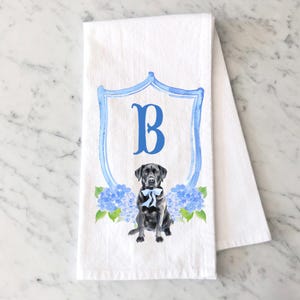 May include: A white kitchen towel featuring a watercolor illustration of a black Labrador dog wearing a light blue bow tie. The dog is centered below a blue crest containing the letter B, flanked by clusters of blue hydrangea flowers and leaves.