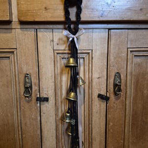May include: A decorative hanging with small brass bells suspended from a black braided cord. A light pink ribbon is tied near the top, and the bells hang against a wooden cabinet.