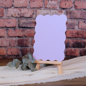 May include: A blank, light purple card with a scalloped edge is displayed on a small wooden easel. The card is sitting on a white fabric surface in front of a brick wall.