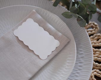 May include: A white place card with scalloped edges sits on a beige linen napkin. The napkin is placed on top of two white plates with a textured design.