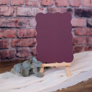 May include: A burgundy card with a scalloped edge is displayed on a small wooden easel. The card is sitting on a white fabric surface with green leaves.