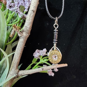 May include: A gold-colored bullet casing necklace with a clear rhinestone in the center. The necklace is on a black cord and has a silver bead accent.