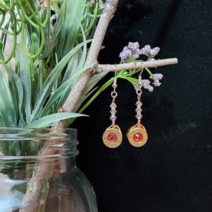 May include: A pair of earrings made from gold-colored bullet casings with red crystal accents. The earrings are suspended from a silver chain with clear crystal beads.