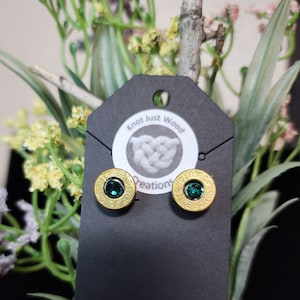 Op de afbeelding: Een paar oorbellen gemaakt van kogelhulzen met groene edelstenen in het midden. De oorbellen worden getoond op een zwarte kaart met de tekst "Knot Just Wood Creations".
