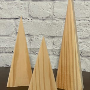 May include: Three wooden triangle shapes in various sizes, standing upright on a brown surface. The triangles are made of light brown wood with visible grain.
