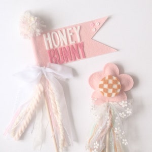 May include: Two decorative wands with pink and white details. One features a pink banner with "HONEY BUNNY" text and a pom-pom. The other is flower-shaped with a checkered centre and a white bow. Both have wooden sticks.