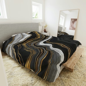 May include: Black and white bedding with gold abstract lines. The bedding is on a bed with a gray headboard and a white rug.