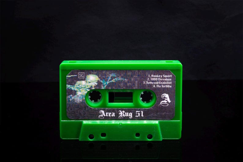 May include: Green cassette tape with a silver label that reads "Area Rug 51". The label also includes the song titles "Honkey Squirt", "1000 Elevators", "Tumcoat Enabilist", and "Flu Tortilla".