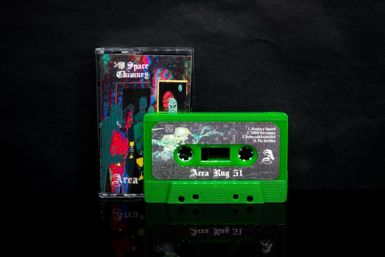 May include: Green cassette tape with the title "Area Rug 51" printed on the front. The cassette tape has a black and white image of a hand holding a microphone. The cassette tape is in a clear plastic case with the title "Space Chimney" printed on the front.