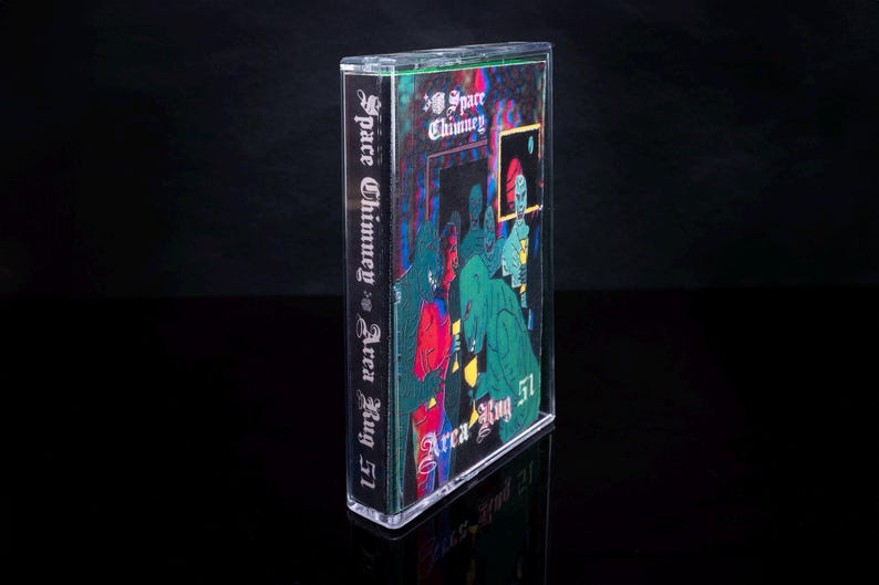 May include: A cassette tape with a black and green illustration of a group of people in front of a building with the text "Space Chimney vs Area Rug 51" on the side.