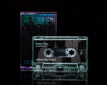 May include: A clear cassette tape with the text "Temporary Creature" and "Colors the Future" printed on it. The cassette tape is in a clear plastic case with a black background.