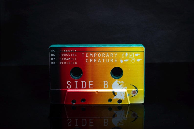 K&ouml;nnte beinhalten: Ein Kassettenband mit einem gelben, orangen und roten Farbverlauf. Das Band ist mit "Temporary Creature" und "Side B" beschriftet. Das Band hat eine schwarze Box mit wei&szlig;em Text, der "05. Blackbox, 06. Crossing, 07. Scramble, 08. Perished" lautet.