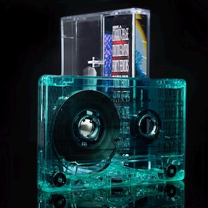 May include: A transparent teal cassette tape with a black reel is displayed in front of a clear cassette case. The cassette tape has a label with text. The background is black.