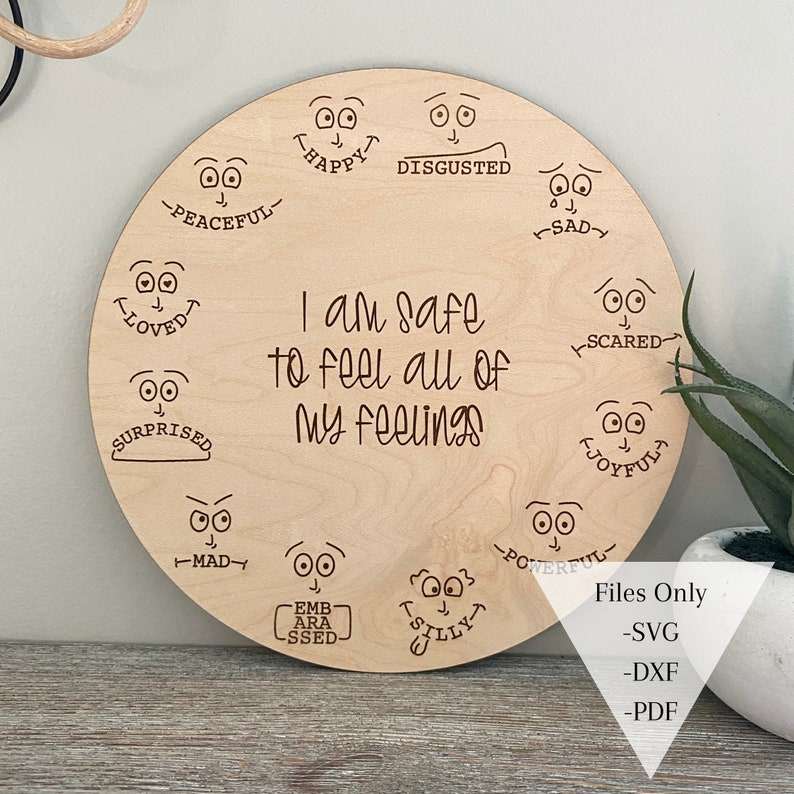 May include: A wooden round sign with the text "I am safe to feel all of my feelings" and various facial expressions around the text. The expressions include happy, sad, scared, surprised, loved, peaceful, disgusted, mad, embarrassed, silly, joyful, and powerful.