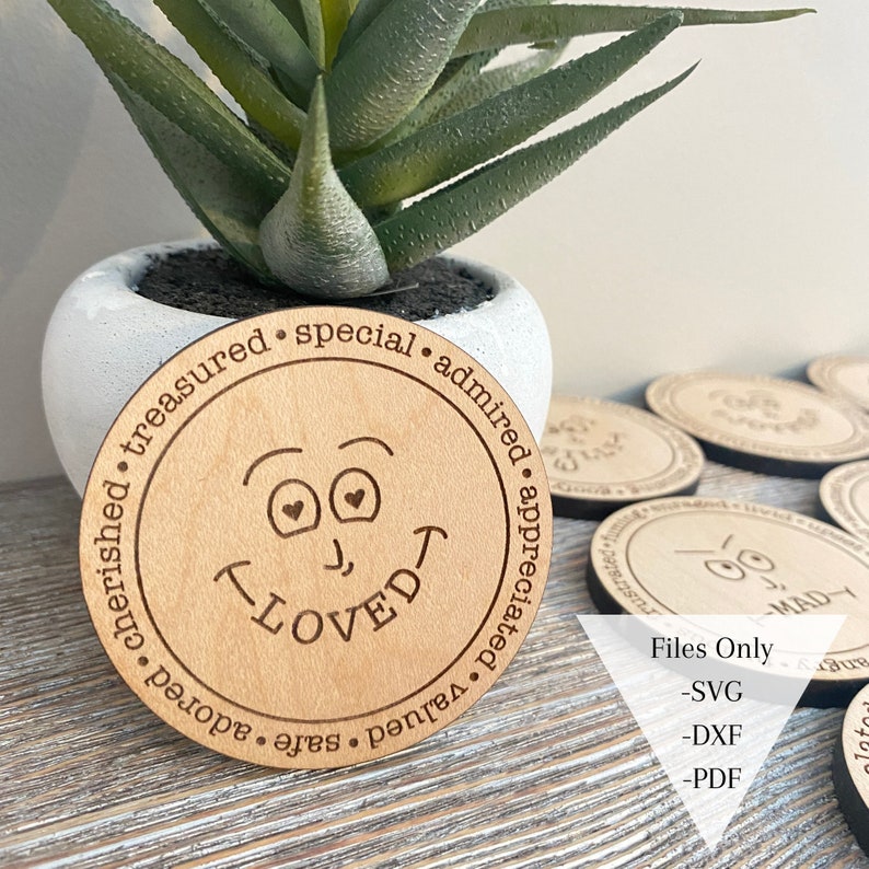 May include: A wooden round coaster with a laser-engraved smiley face with hearts for eyes and the word "LOVED" underneath. The coaster is surrounded by a circle of text that reads "valued, safe, adored, cherished, treasured, special, admired, appreciated".