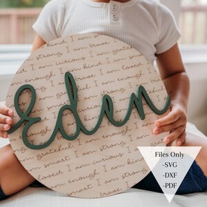 May include: A round wooden sign with the name "Eden" written in large green letters. The sign is covered in a repeating pattern of positive affirmations written in brown ink. The sign is being held by a child.