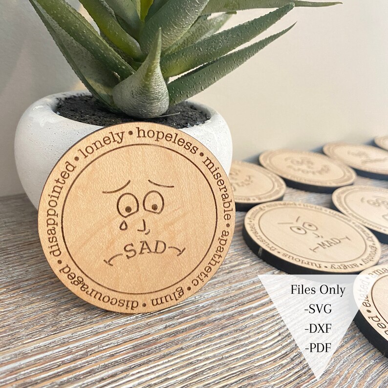 May include: A wooden coaster with a cartoon face and the words "disappointed, lonely, hopeless, miserable, apathetic, glum, discouraged, sad". The coaster is surrounded by other coasters with different cartoon faces.
