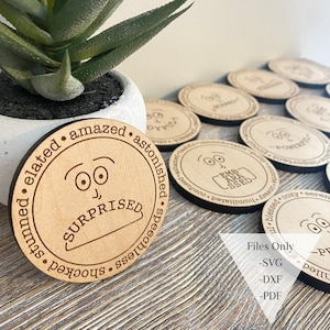May include: A set of wooden coasters with different facial expressions and words describing emotions. The coasters are round and have a natural wood finish. The words on the coasters include "surprised", "elated", "amazed", "astonished", "speechless", "shocked", "stunned", "powerful", "important", "joyful", "comfortable", "humiliated", "confused", "blessed", "cozy", "serene", "uneasy".