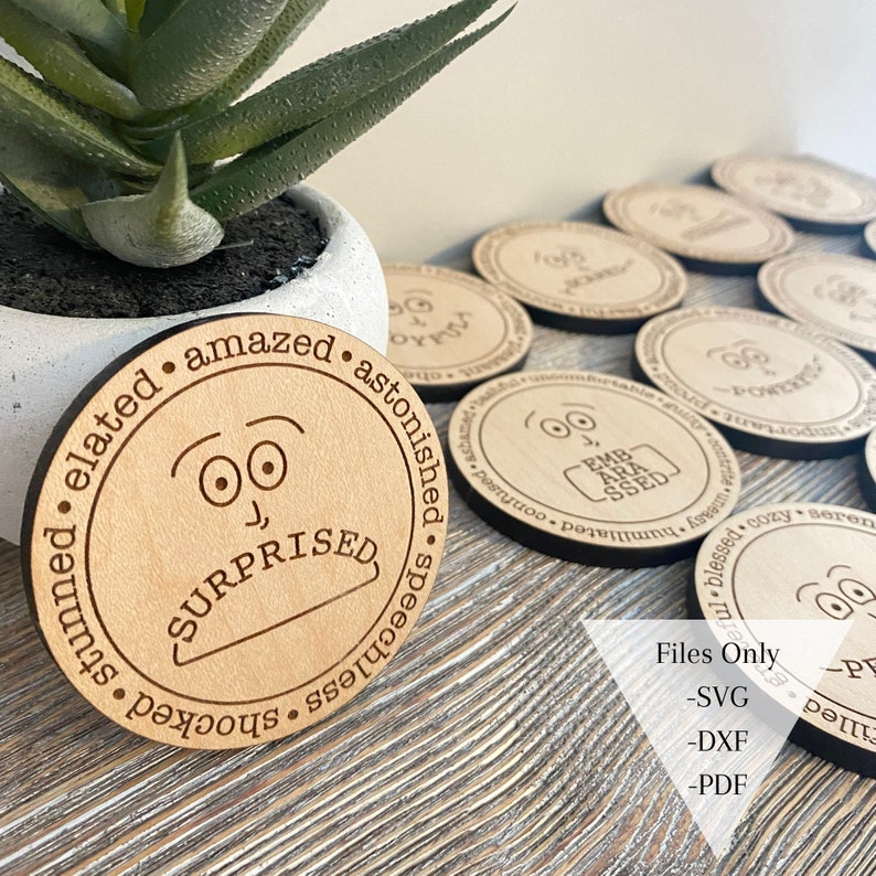 May include: A set of wooden coasters with different facial expressions and words describing emotions. The coasters are round and have a natural wood finish. The words on the coasters include "surprised", "elated", "amazed", "astonished", "speechless", "shocked", "stunned", "powerful", "important", "joyful", "comfortable", "humiliated", "confused", "blessed", "cozy", "serene", "uneasy".