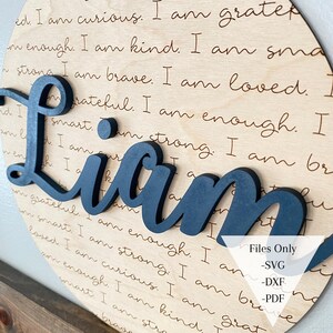 May include: A wooden round sign with the name "Liam" in large, dark blue letters. The sign is decorated with a handwritten message that reads: "I am curious. I am grateful. I am enough. I am kind. I am smart. I am strong. I am brave. I am loved."