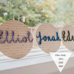 May include: Three round wooden signs with the names "Elliot", "Jonah", and "Eden" written in colorful script. The signs are decorated with a background of brown paper with a repeating pattern of text.