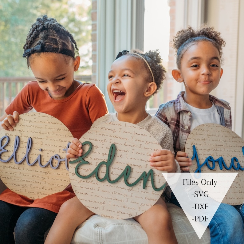 May include: Three children are holding personalized wooden signs with their names, Elliot, Eden, and Jonah, written in cursive. The signs are decorated with a brown and white patterned background and the names are in different colors. The children are smiling and laughing.